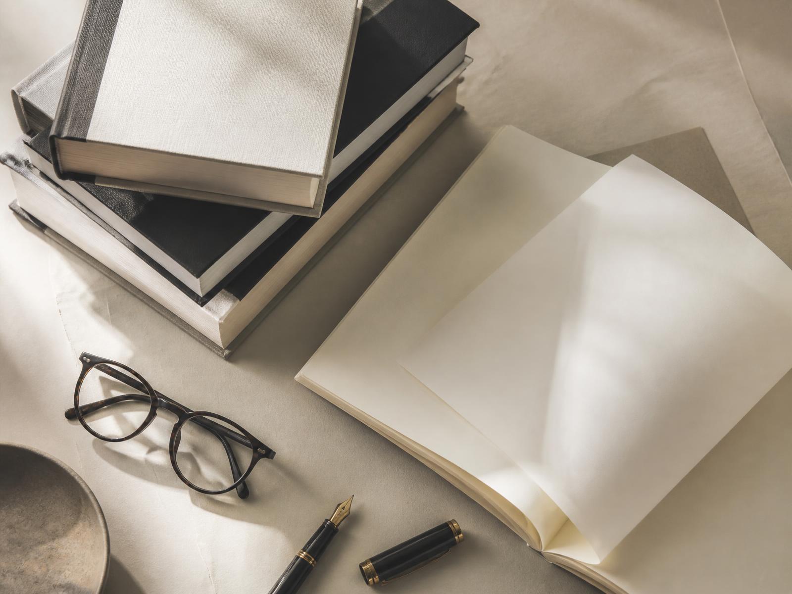 Stack of hardcover books, an open manuscript, reading glasses and a fountain pen on a warm paper-toned desk